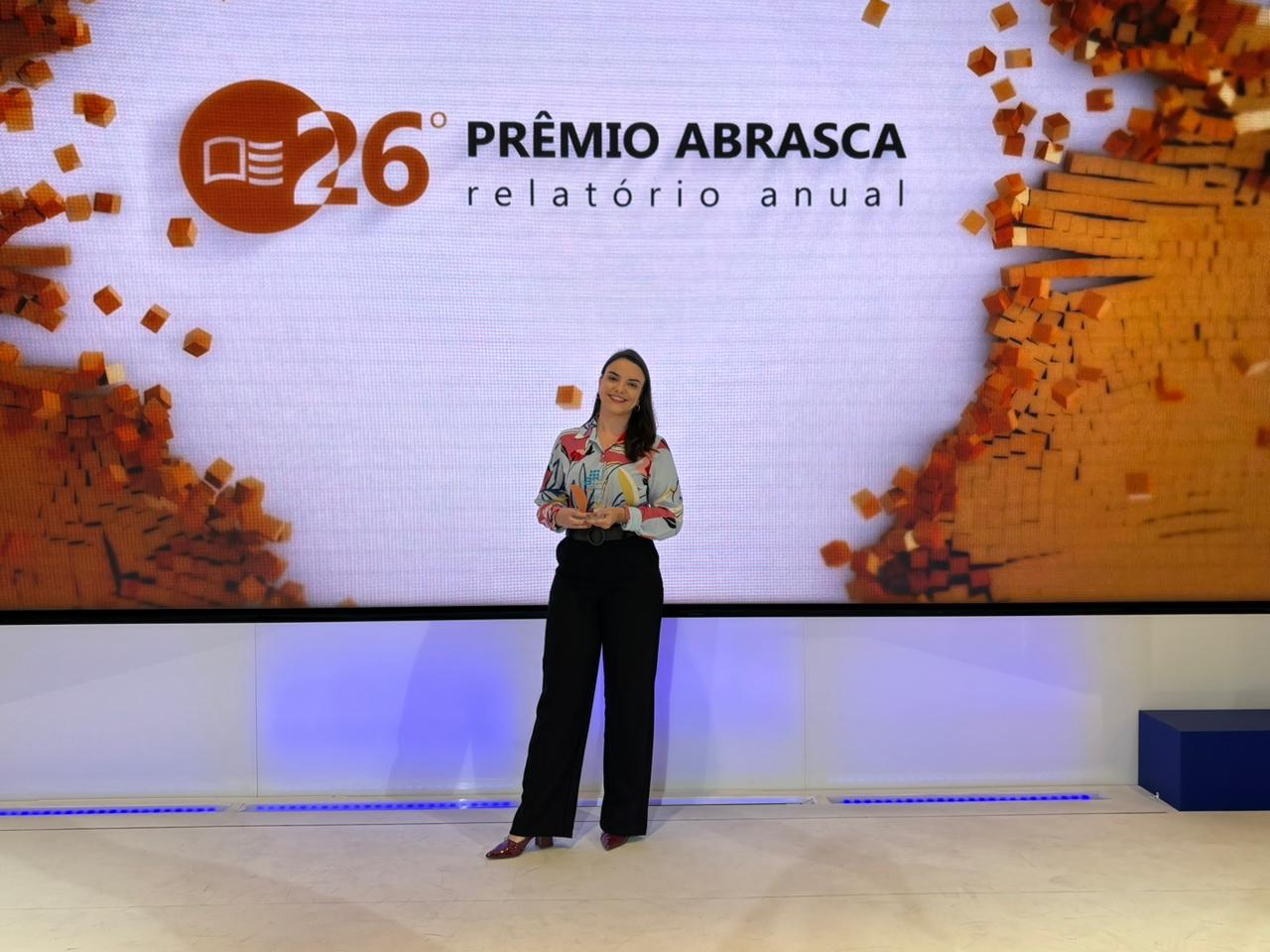 Colaboradora Vittia em frente painel da premiação Abrasca segurando um troféu.